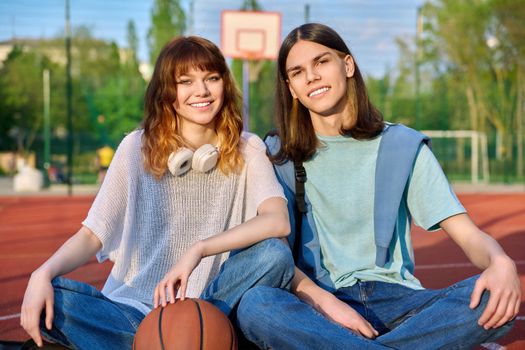 Friends teenagers guy and girl looking at camera, sitting on basketball court