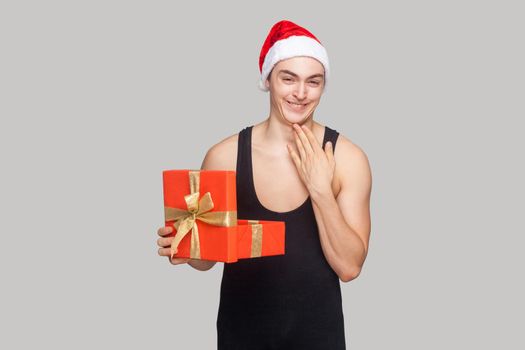 Cunning man holding gift box touching his chin and toothy smiling looking at camera with funny face.