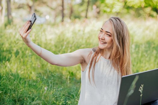 girl sits outdoors and works at a laptop. makes a selfie on the phone. freelance. selfeducation. the concept of remote learning and outdoor work.