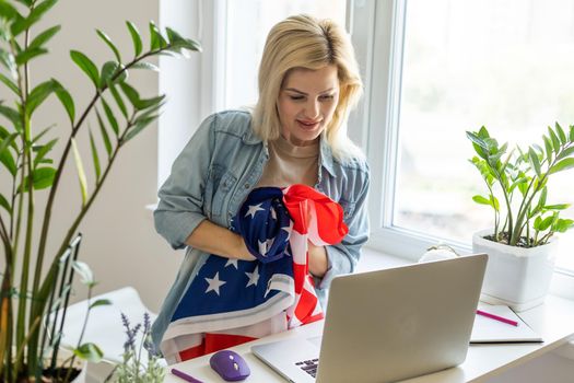 A woman sport fan covered with american flag sits on sofa and watches sport match on laptop in living room and cheers for the american team