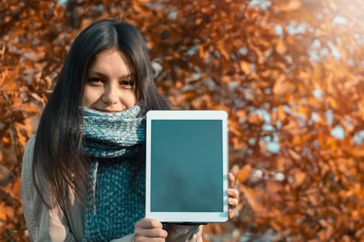 Girl shows the screen of the tablet to the viewer while standing against the backdrop of bright autumn foliage in the park