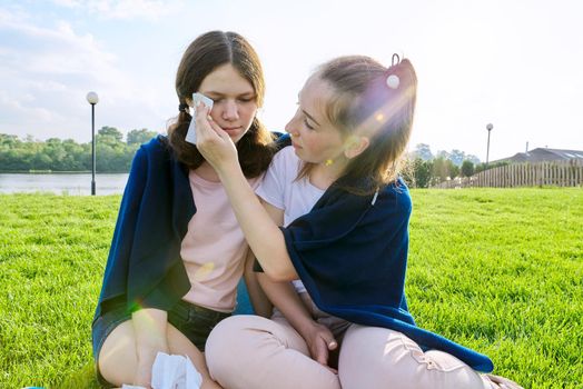 Crying teenager girl and comforting girlfriend, teenagers sitting on the grass in park