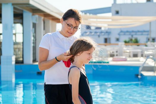 Mom helps daughter child wear swimming cap, outdoor pool background