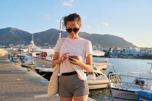 Beautiful teenage girl walking along sea pier with docked yachts, teen with smartphone