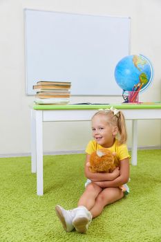 Portrait of cute little latin schoolgirl in classroom. Happy young latin girl wearing casual clothing sitting on a floor holding oy