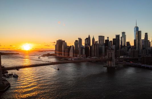 Aerial view of Brooklyn bridge and modern downtown skyscrapers against romantic sunset. Rays of sun reflecting on water surface. Manhattan, New York City, USA
