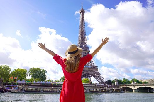 Woman traveler with hat and arms raised on the Seine River in Paris. Back view of fashion woman with open arms in Paris beautiful view with Eiffel Tower on the background.