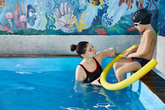 Preschool boy learning to swim in pool with foam noodle with young trainer