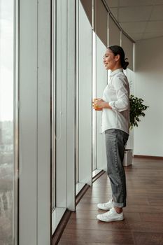 Smiling asian female worker drinking coffee standing near windows at office