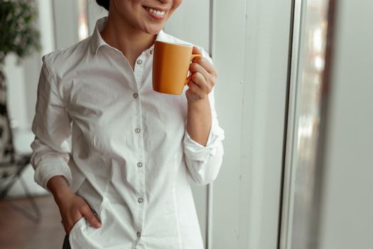 Close up of smiling asian female worker drinking coffee standing near windows at office