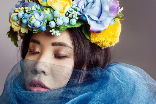 portrait of asian woman with floral hat and blue veil posing with closed eyes on light grey background
