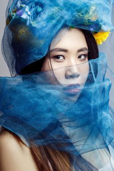 portrait of asian woman with floral hat and blue veil looking at camera on light grey background