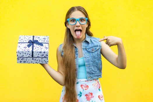 Portrait of a happy hipster smiling girl in dress and blue glasses, holding present box and toothy smile