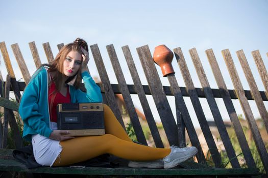 A beautiful country girl in bright clothes sits on a wooden bench with a cassette recorder. Woman in the style of the 90s.