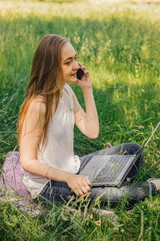 girl sits on the grass and works at a laptop. talking on the phone. freelance. selfeducation. the concept of remote learning and outdoor work.