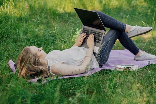 On the banner, a young girl works with a laptop in the fresh air in the park, sitting on the lawn. The concept of remote work. Work as a freelancer. The girl takes courses on a laptop and smiles.