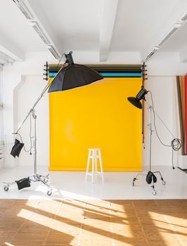 Yellow background roll with a white chair and two professional flash on a c-stands. Sunny daylight photo studio interior with set of professional equipment.