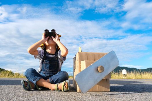 Dreamer little girl looking through binoculars sitting on a lake road beside a cardboard plane against a blue sky background. Childhood dream imagination concept.