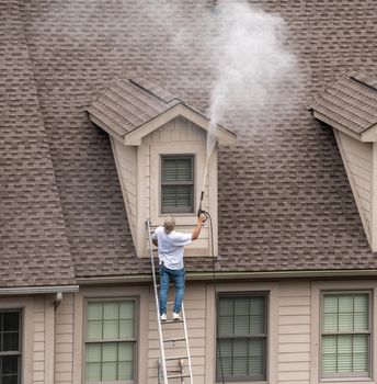 Worker on ladder pressure washing home prior to painting