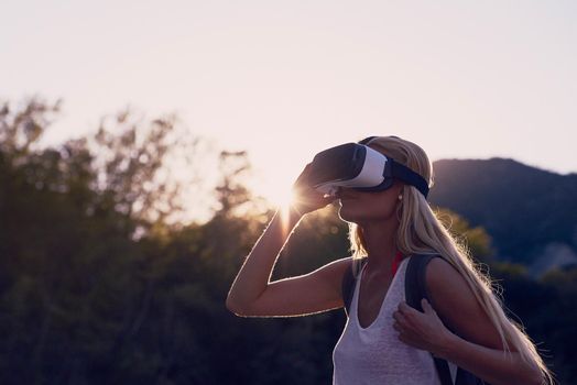 Woman hiking at sunrise using VR goggles to explore the metaverse. Blond woman on holiday hiking in a forest using VR goggles to experience augmented virtual reality