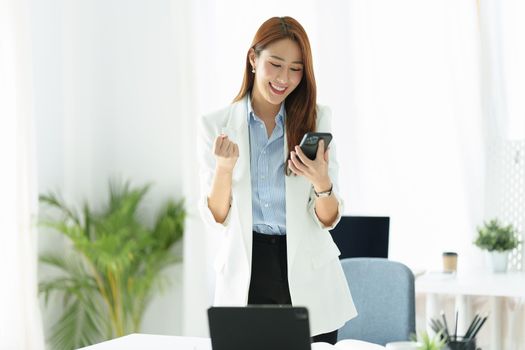 Portrait of a successful Asian businesswoman or business owner expressing excitement and joy using a smartphone mobile in the office