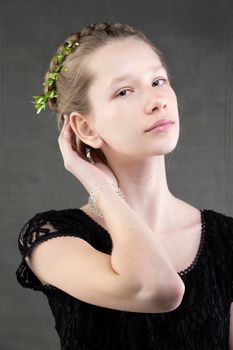 A beautiful teenage girl with a braid with a green twig of a tree woven into it on a gray background. Portrait of a pressed girl with sophisticated features.