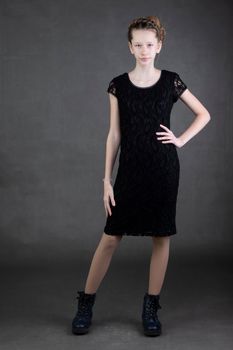 A beautiful thin teenage girl in a black dress and boots is shot on a gray background.