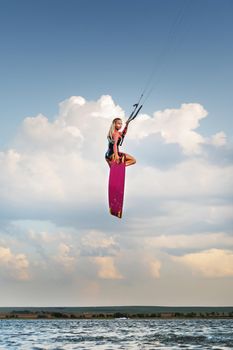 Attractive slender caucasian young woman doing kiteboarding stunt against sunset clouds and beautiful sky. Athlete makes a kitesurfing jump while looking at the camera