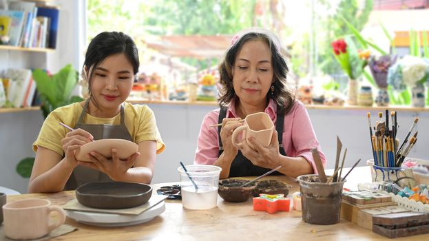 Young woman and mature woman painting pottery in workshop. Indoors lifestyle activity, handicraft, hobbies concept.