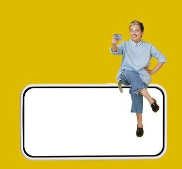 Happy grey haired mature woman sitting on a giant, huge smartphone, in casual blue shirt and jeans with hand raised up in a win, isolated on yellow background