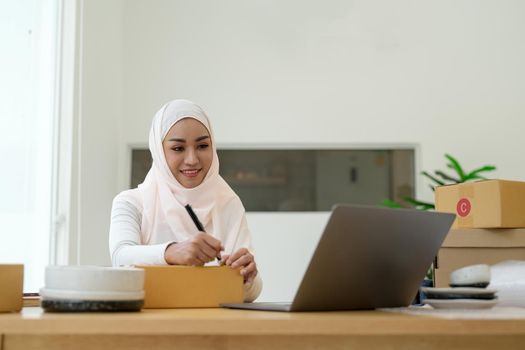 Happy young muslim woman owner writing address on cardboard box, Young Owner Woman Start up for Business Online. People with online shopping SME entrepreneur or freelance working concept.