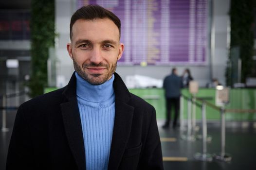 Handsome Middle-Eastern man traveler, passenger stands at flight information board with timetable in international airport arrival departure terminal