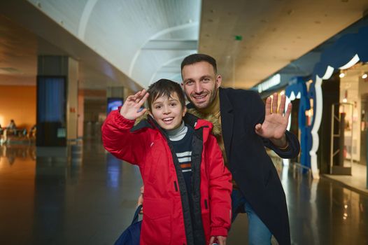 Happy boy and his father, transit passengers smile and wave at camera while walking along duty free shops in international airport departure terminal