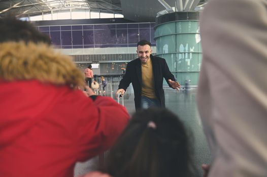 View through people in the foreground of a happy cheerful father running to his family after a long separation in the international airport building
