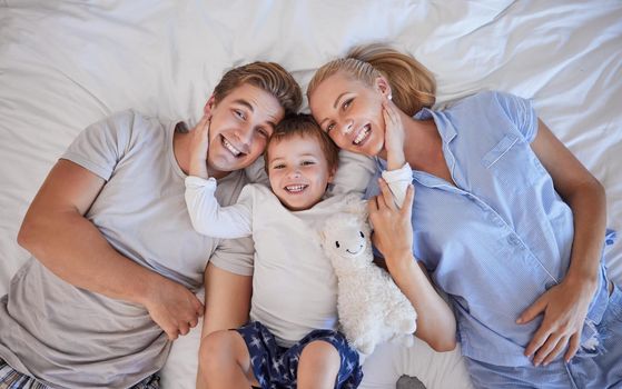 Portrait of adorable little boy with his hands on his parents paces pulling them close while lying on a bed. Cute son lying in between his mother and father, from above. Loving parents bonding with their son