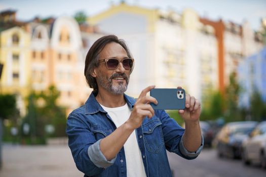Happy middle aged grey bearded man taking pictures using smartphone camera in old town wearing casual. Happy senior man on street of european city with mobile phone. Travel concept