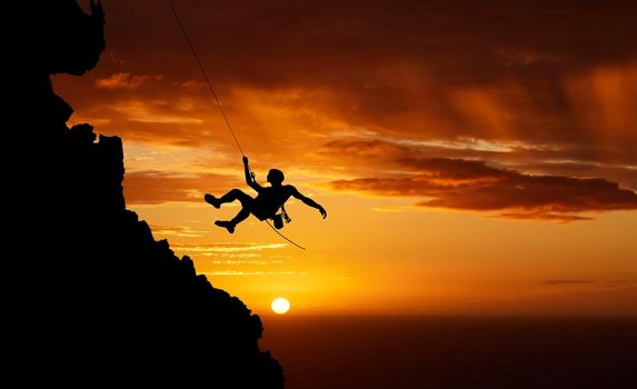 Silhouette of a young male hiker abseiling down the side of a mountain backlit against a sunset sky. Active and sporty man mountain climbing up towards a cliff. Hes a thrill seeking adrenaline junkie