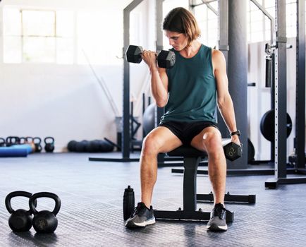 One fit young caucasian man sitting on a bench and doing bicep curls with dumbbells while training in a gym. Focused guy challenging himself by lifting heavy weights to build muscle and endurance during a workout
