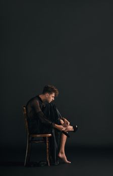 Full body of one handsome caucasian gay man isolated against a black background in a studio and sitting while wearing high heels. Transvestite wearing a dress. Freedom, expression in LGBTQ community