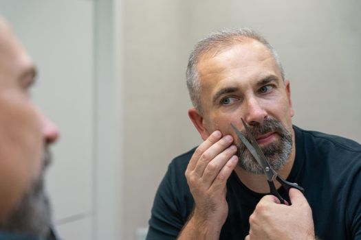 Middle-aged handsome man using scissors to cut his beard a litlie and fix the shape