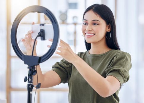 Millennial beauty blogger smiling while adjusting her smart phone on the ring light or pressing record . Beautiful young influencer using a ring light while recording on her mobile phone