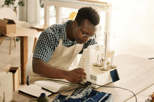 african american tailor sewing a piece of denim. Young designer using his sewing machine. Serious tailor stitching a piece of fabric. Focused seamstress working on his material