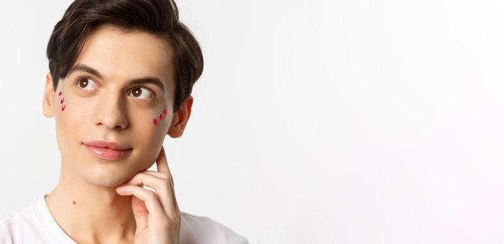 People, lgbtq and beauty concept. Close-up of happy queer man with applied lip gloss and glitter, smiling and looking at upper left corner, standing over white background
