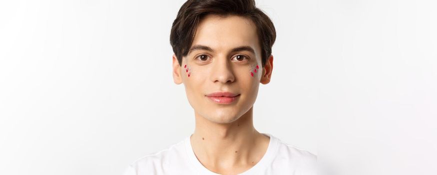 People, lgbtq and beauty concept. Close-up of happy queer guy with applied lip gloss and glitter, smiling and looking at camera, standing over white background