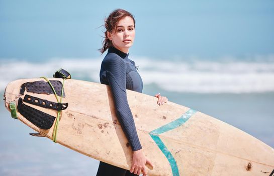 Portrait of beautiful caucasian woman standing alone on a beach, holding a surfboard and wearing a wetsuit before surfing in the sea. Active and athletic woman getting ready to surf waves as a hobby