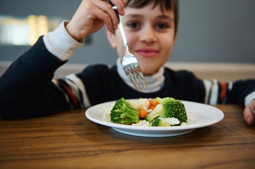 Focus on steamed vegetables, healthy vegan food against a blurred adorable boy holding fork and smiling looking at camera during lunch in cafeteria