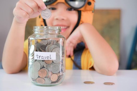 Cute little girl in a pilot hat is putting coins in a clear jar on the table to save for travel. Childhood dream imagination and Travel concepts.