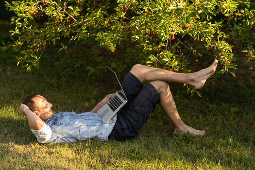 caucasian hispanic man using headphones and sitting on grass working with laptop. Casual man working outdoors