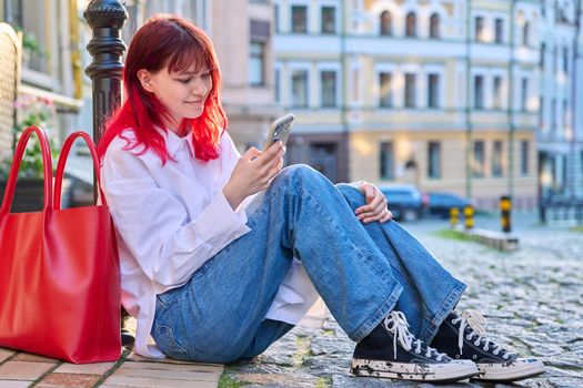 Young fashionable female sitting on the sidewalk with a smartphone