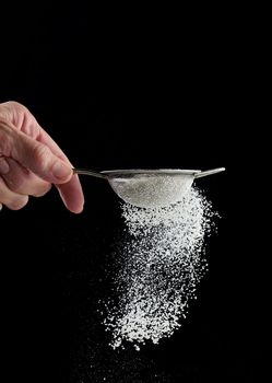 Female hand holds a metal strainer with powdered sugar. Particles fly in different directions 